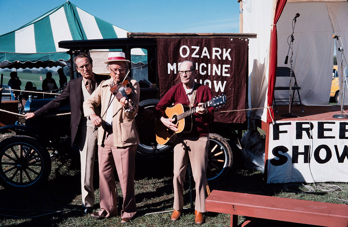 Three elderly men stand in front of an old automobile, with a banner advertising Ozark Medicine Show, among festival tents. One man plays fiddle, another acoustic guitar.