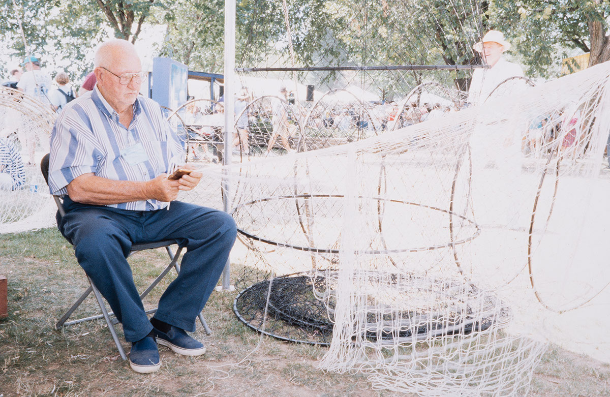 A man, seated, weaves a fishing net from white string.