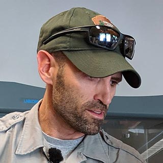 A man wearing a green National Park Service baseball hat with sunglasses resting on the brim. He is wearing a gray shirt and has a beard.