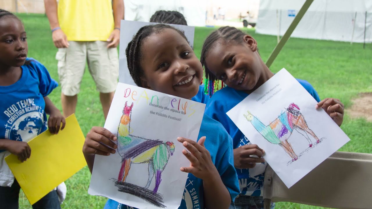 Two young kids hold up coloring pages of alpacas that they have colored in.