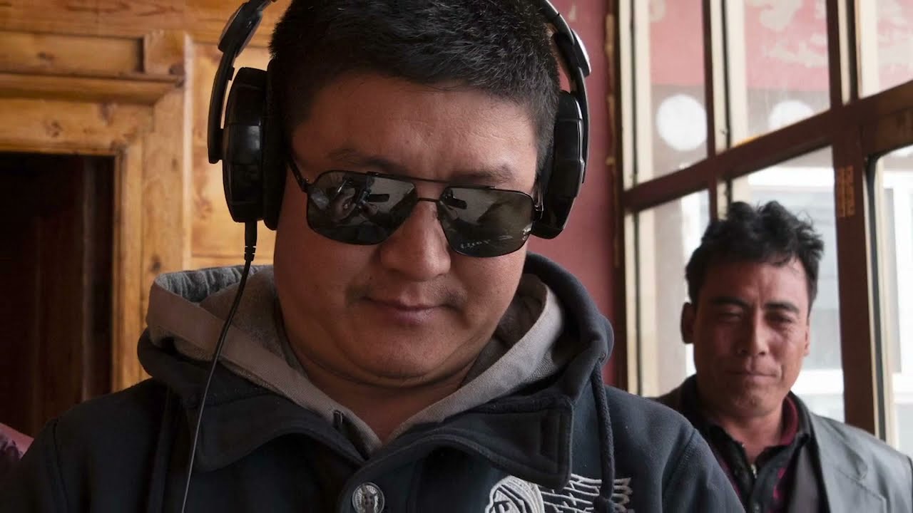 A man watches another man, who wears headphones and dark sunglasses, listening intently.