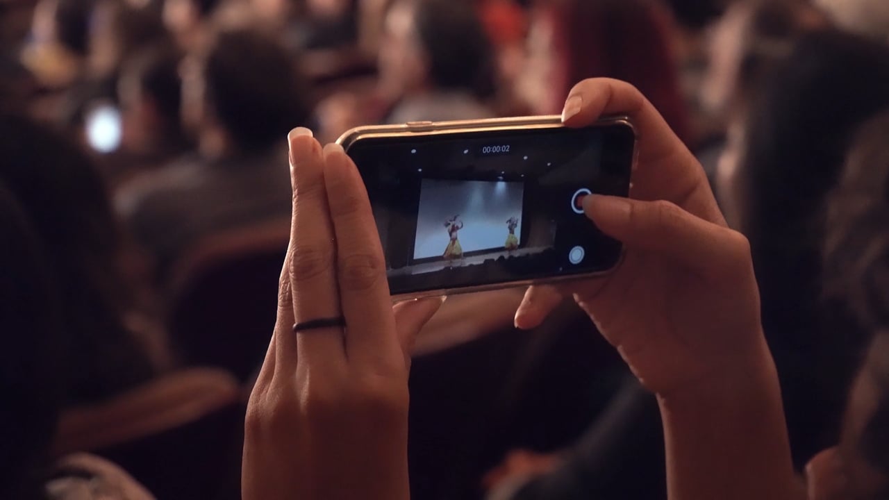 Close-up on two hands filming a dance performance on stage with a smartphone.