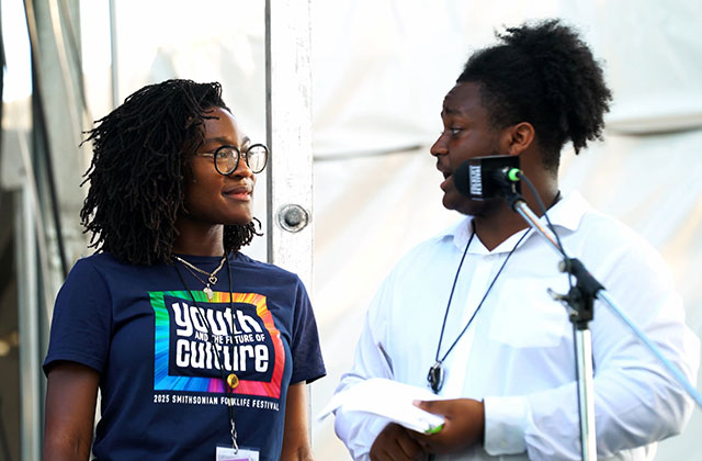 Two young people talk in front of a microphone on stage.